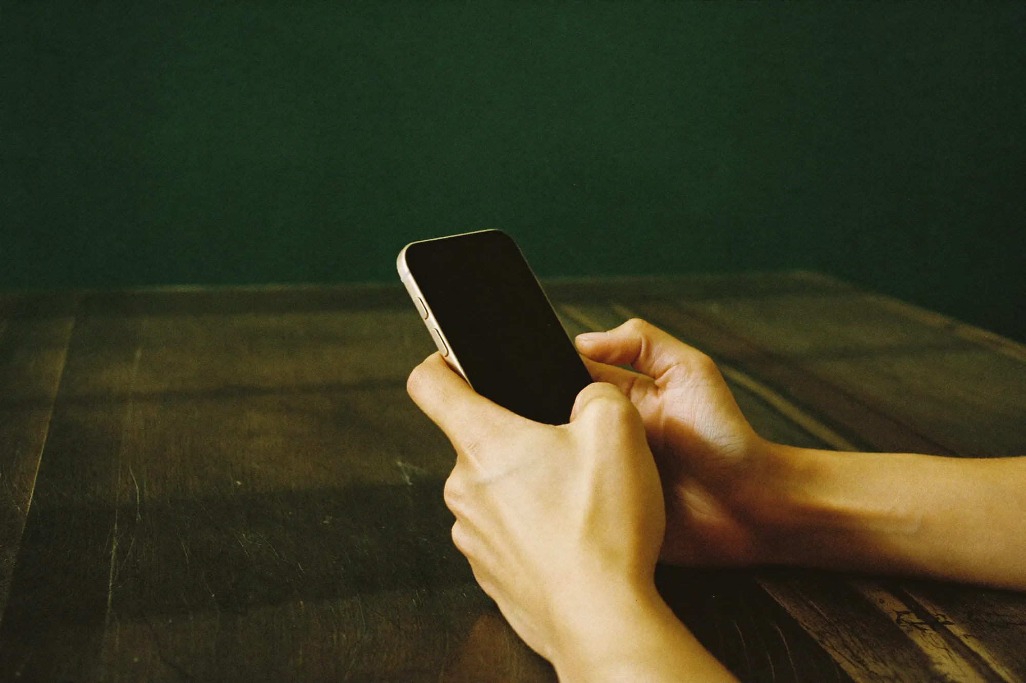 Mãos segurando um celular sobre uma mesa de madeira, parede verde ao fundo